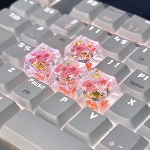 Op de afbeelding: Een close-up van een computertoetsenbord met aangepaste keycaps. De keycaps zijn transparant en bevatten roze bloemen en gouden vlokken. De omliggende toetsen zijn grijs. De "Pause"-toets is zichtbaar.