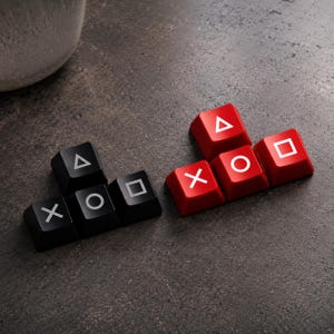 Op de afbeelding: Twee sets PlayStation-controllerknoppen, één zwart en één rood, gerangschikt in een cluster. De zwarte set bevat een X, O, vierkant en driehoek symbool. De rode set heeft dezelfde symbolen. De knoppen liggen op een donkergrijze ondergrond.