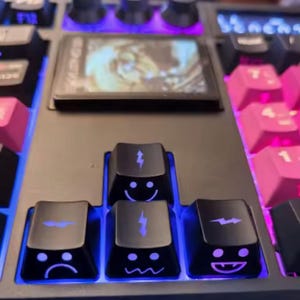 Puede incluir: Un teclado mecánico con teclas negras y rosas. El teclado presenta teclas personalizadas con diseños de caras sonrientes y rayos. Una pequeña pantalla es visible encima de las teclas personalizadas, y el teclado está iluminado con luces azules.