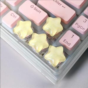 Puede incluir: Primer plano de un teclado de ordenador con teclas rosas y teclas en forma de estrella transparentes. Las teclas en forma de estrella tienen un aspecto amarillo y brillante. El teclado incluye teclas como Ctrl, Shift, Fin y teclas numéricas.