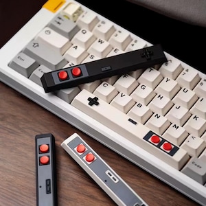 Può includere: Una tastiera meccanica bianca e grigia con tasti in stile retrò e sostituzioni di tasti a forma di controller. I tasti hanno pulsanti rossi e un pad direzionale, evocando un'estetica di gioco vintage.