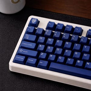 Könnte beinhalten: Eine weiße und blaue mechanische Tastatur mit blauen Tastenkappen. Die Tastatur hat einen weißen Rahmen und dunkelblaue Tasten mit weißer Beschriftung. Die linke obere Taste hat ein Wolfssymbol. Die Tastatur liegt auf einer schwarzen Oberfläche.