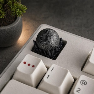 May include: A detailed, metallic Death Star keycap, a spherical design with intricate surface details, sits on a light-colored keyboard. The keycap is surrounded by a black, jagged base. The keyboard keys are white with red and black markings.