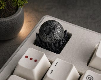 Puede incluir: Una detallada tecla Death Star metálica, un diseño esférico con intrincados detalles de superficie, se encuentra en un teclado de color claro. La tecla está rodeada por una base negra y dentada. Las teclas del teclado son blancas con marcas rojas y negras.