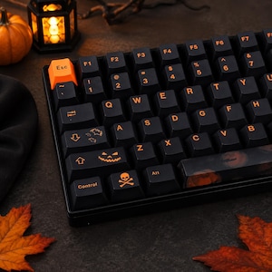 Könnte beinhalten: Eine schwarze mechanische Tastatur mit orangefarbener Beschriftung und individuellen Tastenkappen. Die Escape-Taste ist leuchtend orange, während andere Tasten Symbole wie ein Kürbisgesicht, einen Totenkopf mit gekreuzten Knochen und Fledermäuse zeigen. Die Leertaste hat ein dunkles Kürbismuster.