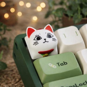 Puede incluir: Una tecla de teclado con forma de gato Maneki-neko blanco, con detalles rojos en las orejas y un collar rojo, colocada en un teclado verde. La tecla tiene una cara detallada con bigotes y ojos negros. La tecla Tab es visible.