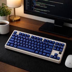 May include: A cream-colored mechanical keyboard with navy blue keycaps sits on a black desk mat. The keyboard is positioned in front of a computer monitor displaying code. A white mouse and a cup of coffee are also on the desk.