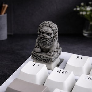 May include: A gray stone Foo Dog statue sits atop a white keyboard key. The keyboard has white keys with black numbers and symbols. The background is dark with a vase of flowers and a pencil holder.