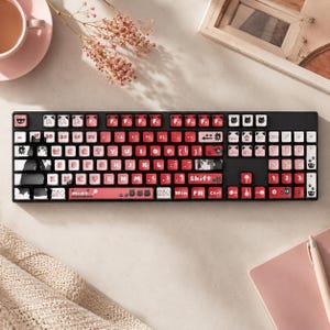 Può includere: Una tastiera meccanica nera con tasti rossi e bianchi. I tasti presentano illustrazioni in stile anime e disegni a tema gatto. La tastiera ha un layout standard con tasti funzione e un tastierino numerico. La tastiera è su una superficie chiara.