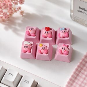 Puede incluir: Un conjunto de seis teclas para teclado mecánico de color rosa claro, cada una con una pequeña figura esculpida de color rosa. Las figuras incluyen variaciones con un corazón rojo, un lazo azul, un gorro de chef blanco, una estrella amarilla y un lazo rosa.
