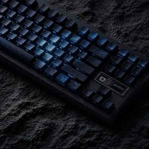 Könnte beinhalten: Eine schwarze mechanische Tastatur mit beleuchteten Tasten. Die Tastenkappen haben ein Galaxie-Design mit blauen und weißen Akzenten. Die Tastatur befindet sich vor einem dunklen, strukturierten Hintergrund.