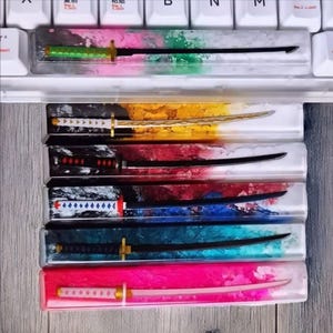 以下が含まれることがあります： 透明なケースに入った、ミニチュアの刀型レターオープナーのコレクション。各オープナーは、緑、金、赤、青、ピンクなど、異なる配色で、ハンドルとブレードのデザインが一致しています。白いキーボードの上に表示されています。