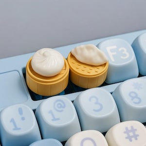 Peut inclure: Gros plan sur un clavier bleu clair avec des touches personnalisées. Deux touches en forme de dim sum, l'une un baozi blanc et l'autre un ravioli blanc, tous deux sur des paniers vapeur en bambou jaunes. D'autres touches sont bleu clair avec des chiffres et symboles blancs.
