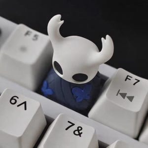 Puede incluir: Una tecla blanca personalizada con un personaje estilizado con cabeza blanca y ojos negros, colocada en un teclado. La tecla tiene una base azul oscuro con detalles de mariposas azules. Las teclas del teclado son blancas con letras negras.