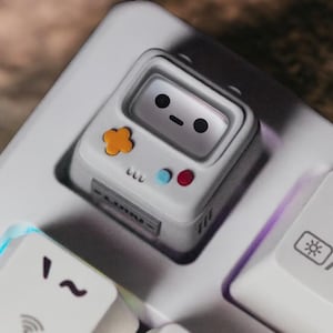 Può includere: Un copritasto bianco per tastiera a forma di console di gioco portatile retrò. Presenta un volto sorridente sullo schermo, un pulsante arancione a forma di fiore e pulsanti circolari blu e rossi. Il copritasto è posizionato su una tastiera bianca.