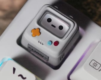 May include: A white keyboard keycap shaped like a retro handheld game console. It features a smiling face on the screen, an orange flower-shaped button, and blue and red circular buttons. The keycap is set on a white keyboard.
