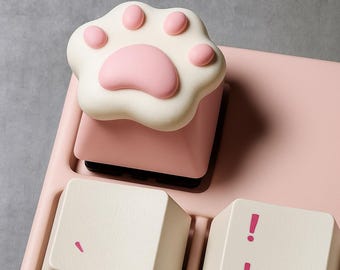 May include: A custom keyboard keycap shaped like a cat paw. The keycap is white with pink paw pads and sits on a pink keyboard. Other keys are off-white with pink accents.