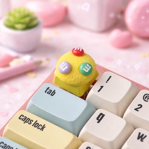 Peut inclure: Un capuchon de touche en forme de cupcake jaune avec des accents rouges, violets et verts est posé sur un clavier. Le clavier a des touches bleu clair, jaunes et blanches. Le capuchon de touche est décoré de ce qui semble être des bonbons.