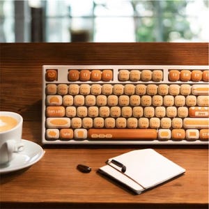 May include: A white and orange mechanical keyboard with custom keycaps featuring cartoon faces. The keyboard sits on a wooden desk next to a cup of coffee and a notepad. The keycaps are a light brown color.
