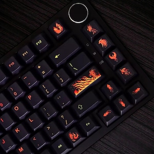 May include: Black mechanical keyboard with custom keycaps featuring orange phoenix designs. The keycaps have a dark background with orange legends and accents. The keyboard has a volume control knob and function keys.