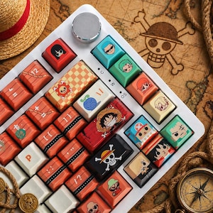 Peut inclure: Un clavier mécanique personnalisé avec des touches représentant des personnages et des symboles de la série animée One Piece. Les touches sont de différentes couleurs et motifs, avec des images de personnages et un Jolly Roger.