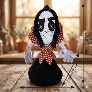 May include: A plush doll featuring a white face with black stitched X-shaped eyes and a black hood. It wears a red and white checkered collar and bodice over a solid black base. Dimensions are 28 cm (11.02 inches) tall, 15 cm (5.91 inches) wide, and 10 cm (3.94 inches) deep.