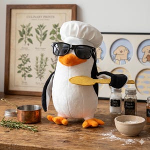 Puede incluir: Un pingüino de peluche con gorro de chef y gafas de sol, sosteniendo una espátula amarilla. El pingüino tiene pico y patas naranjas, con colores blanco y negro. Artículos de cocina, incluyendo especias y una olla de cobre, están en el fondo.