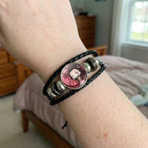 Può includere: Bracciale in pelle intrecciata nera con un ciondolo circolare con un'illustrazione di un personaggio dei cartoni animati. Il ciondolo ha uno sfondo rosa e dettagli argentati. Il bracciale è indossato al polso.