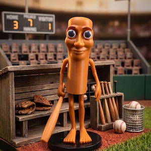 May include: Orange cartoon figure holding a baseball bat, standing on a black base in a miniature baseball stadium. The figure has large eyes and a smiling face. Baseball gloves, bats, and balls are also visible.