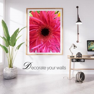 Op de afbeelding: Een ingelijste close-up van een felroze gerbera-madeliefje met een donker centrum. Het kunstwerk wordt getoond op een witte bakstenen muur. De tekst "Decorate your walls" staat eronder.