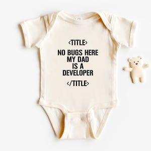 May include: Cream-colored baby onesie with black text that reads "NO BUGS HERE MY DAD IS A DEVELOPER". A small, white teddy bear is next to the onesie. The onesie has short sleeves and snaps at the bottom.