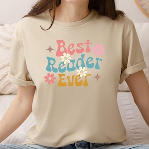 May include: Beige t-shirt with the text "Best Reader Ever" in a retro font. The text is in pink, blue, and yellow with flower and star accents. The shirt has rolled-up sleeves.