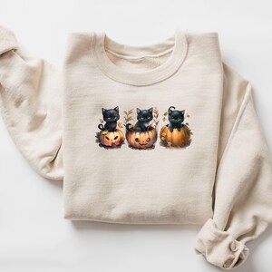 May include: A cream-colored sweatshirt featuring a watercolor illustration of three black kittens sitting in carved orange pumpkins. The pumpkins have jack-o'-lantern faces and are surrounded by autumn leaves, creating a festive Halloween theme.