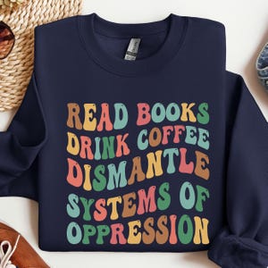 May include: Navy blue sweatshirt with the text "READ BOOKS DRINK COFFEE DISMANTLE SYSTEMS OF OPPRESSION" in colorful, retro-style lettering. The sweatshirt is folded, showing the front design.
