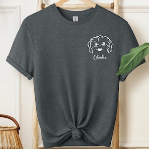 May include: Dark gray t-shirt featuring a white line drawing of a dog's face with the name "Charlie" below it. The t-shirt is knotted at the waist and hangs on a wooden hanger.