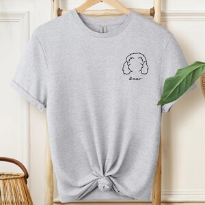 May include: A light gray t-shirt with a knotted front. The shirt features a black line drawing of a dog's face and the name "Bear" in a handwritten font. The shirt is hanging on a wooden rack.