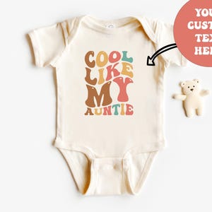 May include: A cream-colored short-sleeve baby bodysuit featuring the text COOL LIKE MY AUNTIE in a multi-colored retro font. A small white bear figurine is positioned to the right, and a coral circle with the text YOUR CUSTOM TEXT HERE.