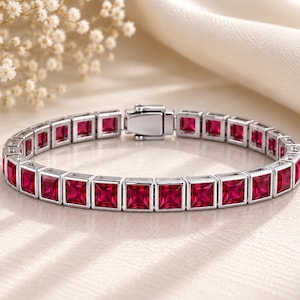 Puede incluir: Una pulsera tipo tenis de tono plateado con una hilera continua de gemas rojas vibrantes de corte cuadrado, engastadas en marcos metálicos individuales. La pulsera incluye un mecanismo de cierre de caja integrado y seguro.