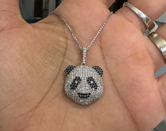 Peut inclure: Un collier en argent avec un pendentif panda. Le pendentif panda est incrusté de petites pierres blanches scintillantes, avec des accents noirs pour les yeux, les oreilles et le nez. Le collier est présenté sur une main.
