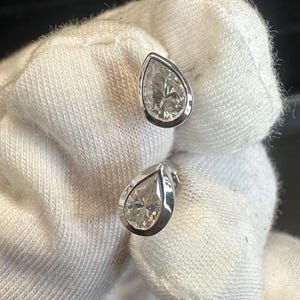 May include: A pair of pear-shaped diamond stud earrings. Each earring features a clear, sparkling diamond set in a silver or white gold bezel. The earrings are displayed against a white background.