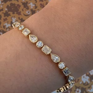 Puede incluir: Una pulsera dorada con una serie de diamantes engarzados en bisel de varias formas, incluyendo redondos, pera y esmeralda. Los diamantes son claros y brillantes, engarzados en una cálida banda dorada. Una pieza de joyería fina.