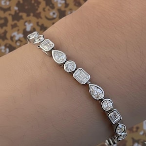 May include: A silver bracelet with a series of alternating diamond shapes, including round, pear, and emerald cuts. The bracelet is set with multiple clear, sparkling gemstones. The jewellery is displayed on a person's wrist.