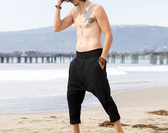 May include: A man wearing black, loose-fitting pants with a drawstring waist. He is standing on a sandy beach with a pier in the background.