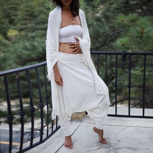 May include: A woman wearing a white, loose-fitting,  cropped top and white, flowing pants with a draped, white, sheer fabric over her shoulders. She is standing on a balcony with a black railing.