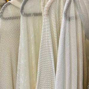 May include: Five white crocheted sweaters hanging on hangers. The sweaters are all different textures and have a loose, flowy fit.
