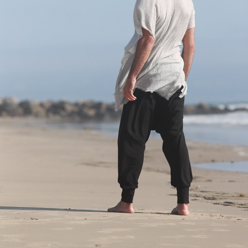 May include: A person wearing a white, loose-fitting shirt and black pants stands on a sandy beach.