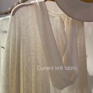 May include: A white knit hooded cardigan with a loose fit. The fabric is lightweight and has a delicate texture. The cardigan is hanging on a wooden hanger.
