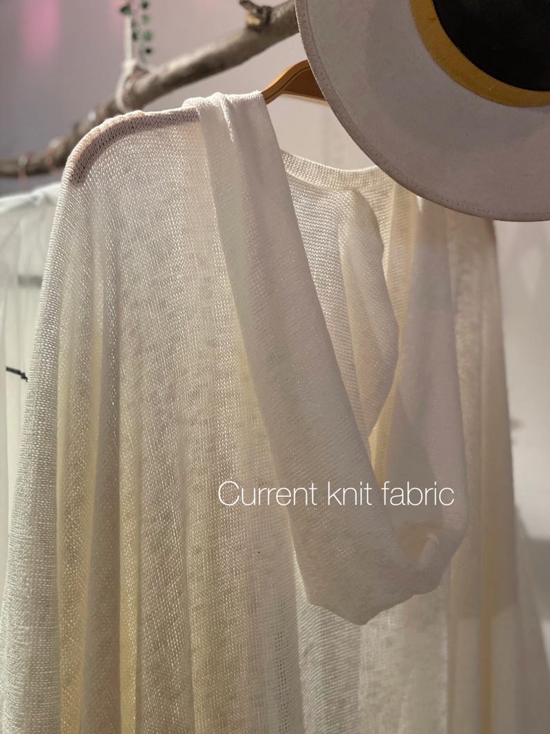 May include: A white knit hooded cardigan with a loose fit. The fabric is lightweight and has a delicate texture. The cardigan is hanging on a wooden hanger.