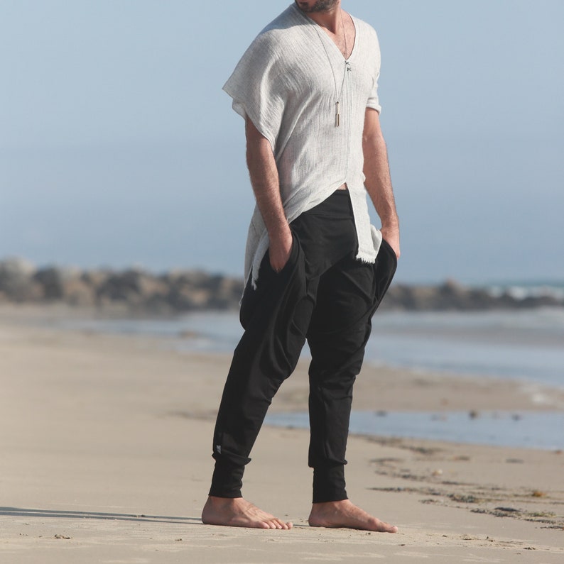 May include: A person wearing a white, loose-fitting shirt with a long pendant necklace and black pants. The person is standing on a sandy beach.
