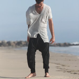 May include: A man wearing a white, loose-fitting shirt with a V-neck and black, loose-fitting pants. He is standing on a sandy beach.